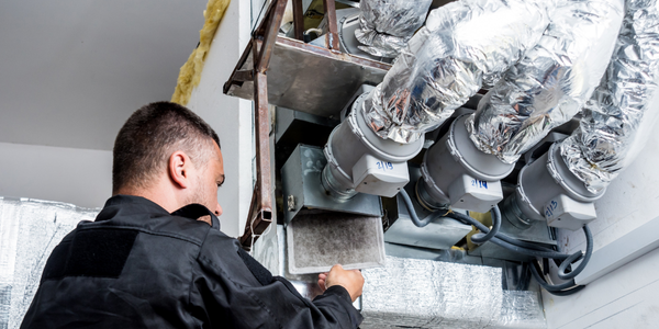 A technician is conducting a thorough inspection of HVAC ductwork in a building. He is carefully assessing a filter, surrounded by insulated ducts and ventilation equipment. The setting appears industrial or commercial, with a focus on air quality management.