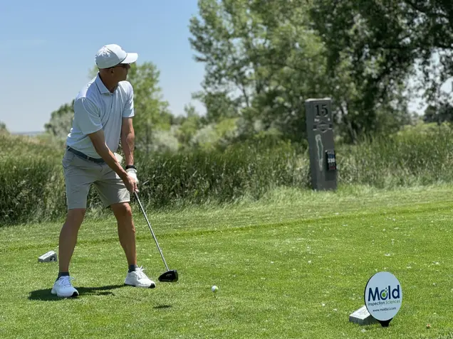 A golfer in a white cap and shirt prepares to swing on the golf course, where a Mold Inspection Sciences sign nestles in the grass. Trees frame the scene, with a sign indicating it's the 15th hole visible in the background.