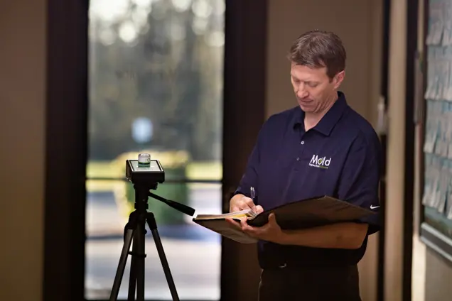 A man in a navy blue shirt is standing indoors, sampling data as he writes on a clipboard. A camera on a tripod is positioned in front of him, possibly recording the testing process. The blurred background shows a door leading outside with sunlight streaming in.