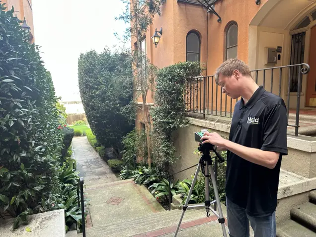 A man in a black Meld polo shirt adjusts a camera on a tripod outside, standing on steps beside an orange building—a scene perfect for mold assessment or sampling near the green garden path.