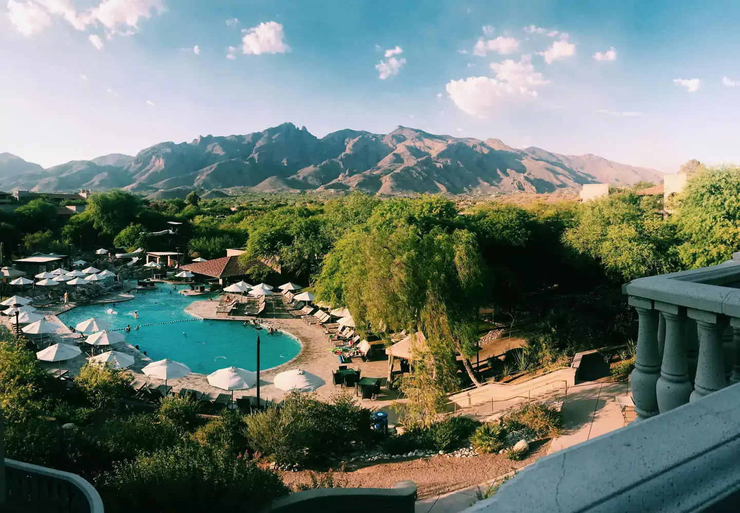 A scenic view of a resort pool area with umbrellas and lounge chairs, surrounded by lush greenery, stands out during a meticulous assessment of the venues offerings. In the background, a mountain range under a blue sky with scattered clouds creates a stunning backdrop.